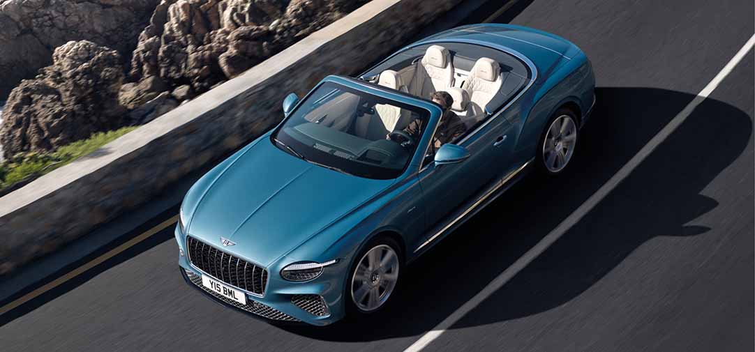 Overhead view of the Bentley Continental GTC Azure convertible cruising along a coastal road with the top down, revealing its luxurious white interior and ocean-inspired blue exterior.
