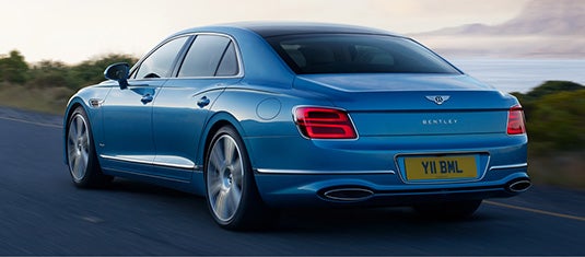 Rear view of the Bentley Flying Spur Azure driving along a coastal road, revealing its sculpted rear design, LED tail lights, and dual exhaust finishers beneath the Bentley badge.