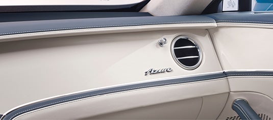 Close-up of the Bentley Flying Spur Azure dashboard, showcasing the Azure emblem, round air vent, and stitched leather with chrome accents.