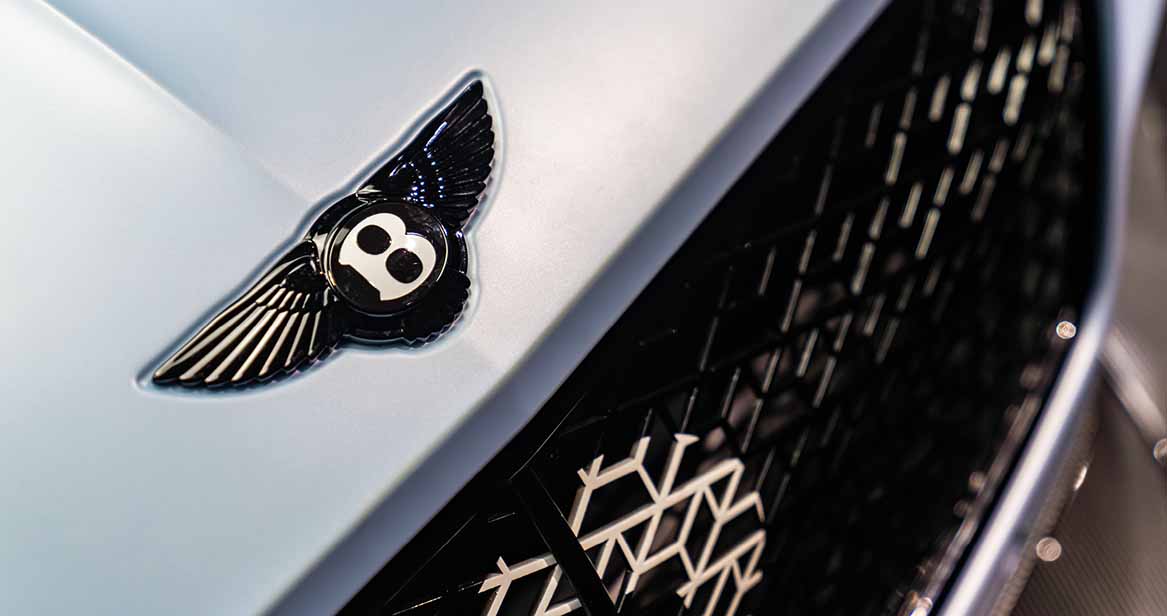 Macro shot of Bentley’s blacked-out winged 'B' emblem on a satin-finish hood, with grille detail in background representing bespoke British automotive craftsmanship.